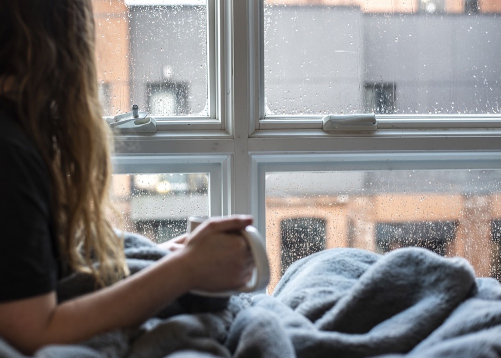 A woman wrapped in a blanket looking out a rain-streaked window with a warm drink